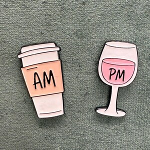 Könnte beinhalten: Zwei Emaille-Pins, einer zeigt eine Kaffeetasse mit dem Text "AM" und der andere ein Weinglas mit dem Text "PM".