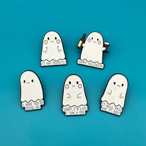 Może przedstawiać: Pięć białych emaliowanych spinek z duchami z różnymi powiedzeniami. Spinki przedstawiają uroczy projekt ducha z kreskówek. Powiedzenia na spinkach to "Ma kieszenie!" "Szczęśliwego Halloweine" "Mam nadzieję, że dobrze się masz!" "Przepraszam, spóźniłem się, widziałem psa" i "To minie i tak się dzieje."