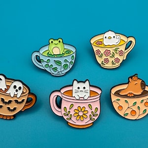 Könnte beinhalten: Set aus fünf Emaille-Ansteckern mit niedlichen Charakteren in Teetassen. Enthält einen Frosch, einen Vogel, zwei Geister, eine Katze und ein Capybara. Die Teetassen sind mit Blumen- und Blattmotiven verziert.