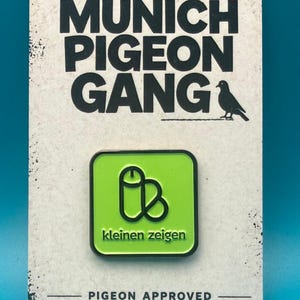 Könnte beinhalten: Ein quadratischer Emaille-Pin mit lindgrünem Hintergrund und einer schwarzen Umrandung in phallischer Form, der den schwarzen Schriftzug 'kleinen zeigen' trägt. Der Pin ist auf einer weißen Karte mit 'MUNICH PIGEON GANG' und 'PIGEON APPROVED' befestigt.