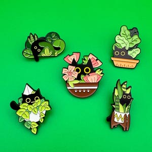 May include: Five enamel pins featuring black cats in different plant pots. The cats are all looking at the viewer with big eyes. The plants are a variety of green and pink.