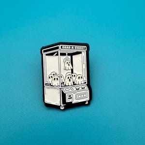 Peut inclure: Épingle en émail noir et blanc représentant une machine à griffes avec le texte "GRAB-A-CHOST". La machine contient plusieurs fantômes blancs. L'épingle est posée sur un fond turquoise.