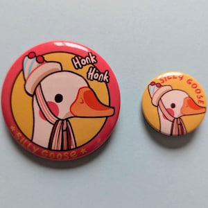Può includere: Due spille rotonde raffiguranti un'oca dei cartoni animati con un cappello e una sciarpa a righe. La spilla più grande ha un bordo rosa, uno sfondo giallo e il testo Honk Honk e SILLY GOOSE. La più piccola ha uno sfondo giallo e SILLY GOOSE.