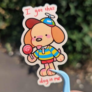 May include: A die-cut sticker featuring a cartoon dog wearing a blue and yellow striped shirt, red shorts, and a propeller cap. The dog holds a pink lollipop and has the red text "I got that dog in me" printed above and below the character illustration.
