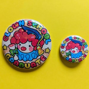 May include: Two round, colorful buttons with a clown theme on a yellow background. The larger button features a clown with red hair, a blue collar, and the words "Clown School Graduate." The smaller button has the same design.