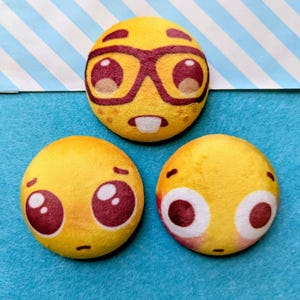 May include: Three yellow plush emoji-style circles with different facial expressions. One has glasses and a buck tooth, another has sad eyes, and the third has wide eyes. The background is blue with a white and blue striped pattern.