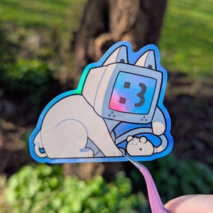May include: A holographic sticker depicting a cat with a retro computer monitor head. The monitor shows the number 3 and a heart symbol. The cat is light grey with blue accents and a small, white creature.