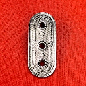 May include: An elongated, oval-shaped silver-toned metal button featuring three centered circular holes. The surface is decorated with a fine, textured cross-hatch pattern, engraved geometric borders, and small star-like motifs between the holes.