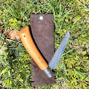 May include: A folding saw with a wooden handle and a stainless steel blade, partially extended from a brown leather sheath. The handle has the word "OPINEL" printed on it. The saw is resting on a bed of green grass.