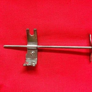 May include: A metal sewing machine part with a silver rod and a U-shaped piece. The part is attached to a rectangular piece with a small screw. The background is a solid red color.
