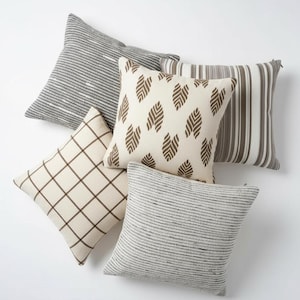 May include: An assortment of decorative throw pillows in various patterns and colors. Pillows include striped, grid, and leaf designs in neutral tones of gray, brown, and cream. The pillows are square and arranged on a white surface.