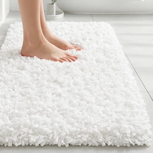 以下が含まれることがあります： 白く長方形のバスマットで、毛足の長い質感のある表面です。マットは柔らかい素材でできており、タイルの床に置かれています。画像には、マットの上に立っている人の足が写っています。
