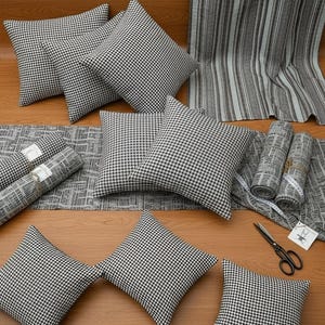 Puede incluir: Una colección de artículos de decoración para el hogar, que incluye cojines, rollos de tela y una cortina, todos con un patrón de cuadros blancos y negros. Los artículos están dispuestos sobre una superficie de madera, con unas tijeras y una cinta métrica.