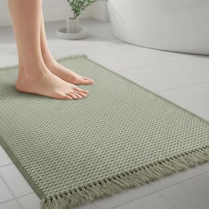 May include: A rectangular sage green bath mat with a textured, waffle-weave pattern and fringed edges. The mat is placed on a tiled floor, with a person's feet standing on it. The mat is designed for the bathroom.