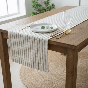 Puede incluir: Una mesa de comedor de madera con un camino de mesa a rayas, un plato blanco con vegetación y cubiertos dorados. Una copa de vino transparente está al lado del plato. Una alfombra redonda de yute está debajo de la mesa.