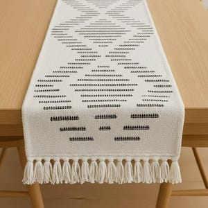 Op de afbeelding: Crèmekleurige tafelloper met een geometrisch patroon in zwart en wit. De loper heeft een reeks horizontale lijnen en ruitvormen, met een franje aan de uiteinden. De loper ligt op een houten tafel.