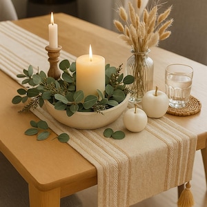 Op de afbeelding: Een tafeldecoratie met een brandende kaars en groen in een witte kom, naast een beige gestreepte tafelloper met kwastjes. Twee witte pompoenen en een vaas met droogbloemen creëren een warme, herfstige sfeer.