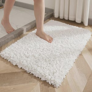Op de afbeelding: Een rechthoekige witte badmat met een getextureerd, gerimpeld oppervlak. De mat ligt op een houten vloer en een persoon stapt erop. De mat ziet er zacht en pluche uit, ontworpen voor gebruik in de badkamer.