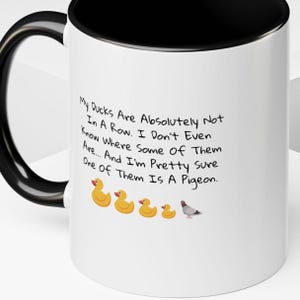 Puede incluir: Taza de cerámica blanca con borde y asa negros. La taza tiene una frase humorística sobre patos y una paloma impresa en ella. El texto dice: "Mis patos no están absolutamente en fila. Ni siquiera sé dónde están algunos de ellos.... Y estoy bastante seguro de que uno de ellos es una paloma."