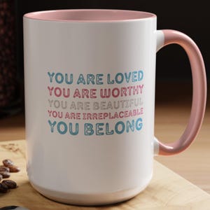 Könnte beinhalten: Weißer Keramikbecher mit rosa Innenraum und Henkel. Der Becher trägt den Text "YOU ARE LOVED, YOU ARE WORTHY, YOU ARE BEAUTIFUL, YOU ARE IRREPLACEABLE, YOU BELONG" in blauen und rosa Buchstaben. Der Becher steht auf einer Holzoberfläche.
