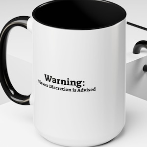 Può includere: Tazza in ceramica bianca con bordo e manico neri. La tazza ha la scritta "Avvertenza: Si consiglia la discrezione dello spettatore" stampata su di essa.