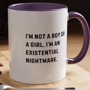 Puede incluir: Taza de cerámica blanca con interior y asa morados. La taza presenta el texto negro «I'M NOT A BOY OR A GIRL. I'M AN EXISTENTIAL NIGHTMARE» centrado en la parte delantera. La taza está sobre una superficie de madera.