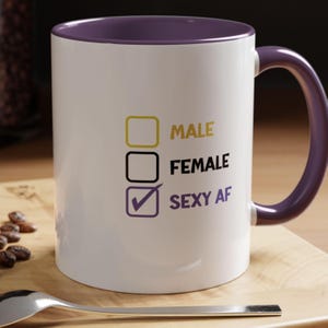 Puede incluir: Taza de cerámica blanca con interior y asa morados. La taza presenta los textos "MALE", "FEMALE" y "SEXY AF" con casillas de verificación. Granos de café y una cuchara en primer plano.