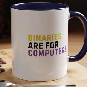 May include: White ceramic mug with a navy blue handle and rim. The mug features the text "BINARIES ARE FOR COMPUTERS" in yellow, black, and purple lettering. The mug is sitting on a wooden surface.