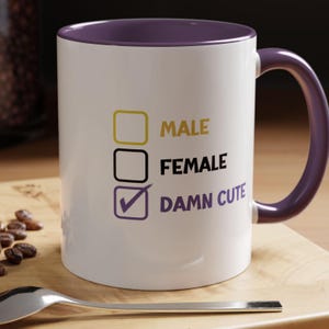 Puede incluir: Taza de cerámica blanca con interior y asa morados. La taza presenta los textos "MALE", "FEMALE" y "DAMN CUTE" con casillas de verificación. La taza está sobre una superficie de madera.