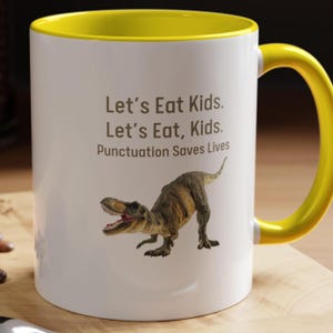 Puede incluir: Taza de café de cerámica blanca con borde y asa amarillos. La taza presenta una ilustración de dinosaurio marrón y el texto "Let's Eat Kids. Let's Eat, Kids. Punctuation Saves Lives."