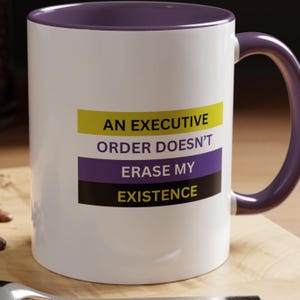 Puede incluir: Taza de cerámica blanca con borde y asa morados. La taza tiene un gráfico con el texto "An Executive Order Doesn't Erase My Existence" en texto negro sobre un fondo a rayas amarillo, morado y negro.