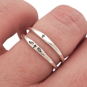May include: Two silver rings, each with a different engraved design. One ring has a cross, the other a cross and wings. The rings are shown on a finger against a white backdrop.