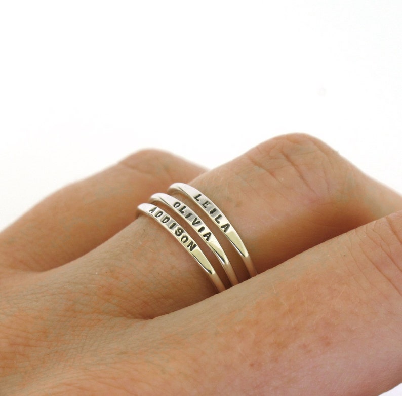 Close up picture of a finger wearing 3 very tiny sterling silver rings.  Each ring is hand stamped with a different name.  The letters are 1mm tall.