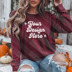 May include: A maroon long-sleeved sweatshirt with the text "Your Design Here" in white retro-style lettering. The sweatshirt is worn by a person with long blonde hair and is sitting on a wooden surface with pumpkins in the background.