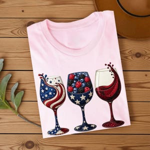 May include: A light pink t-shirt featuring a graphic of three wine glasses. The first glass has a red, white, and blue star and stripe pattern. The second glass is filled with red and blue berries. The third glass contains red wine.