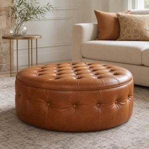 May include: A round, tan-colored leather ottoman featuring a tufted button design on the top surface and along the sides. The piece has a smooth, uniform texture and a cylindrical shape, providing a low-profile seating or footrest surface.