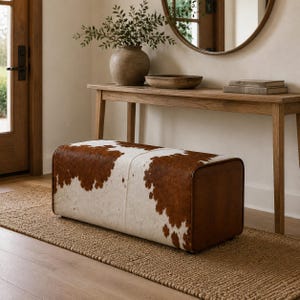 Peut inclure: Un pouf rectangulaire recouvert de peau de vache marron et blanche avec un panneau latéral en cuir marron lisse. Le meuble présente un design bas aux bords arrondis et une construction sans couture, mettant en valeur la texture naturelle.