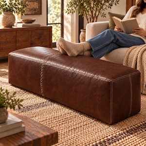 Pode incluir: Um otomano retangular de couro castanho com uma superfície lisa e uma costura vertical proeminente com detalhes decorativos em ponto cruz. O otomano tem um design estruturado de perfil baixo com bordas limpas e costuras visíveis.
