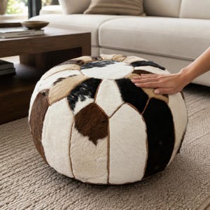 Peut inclure: Un pouf rond composé de panneaux de peau de vache cousus dans un motif patchwork. La surface présente un mélange de fourrure blanche, noire et de différentes nuances de brun, disposées en un motif géométrique avec des coutures visibles.