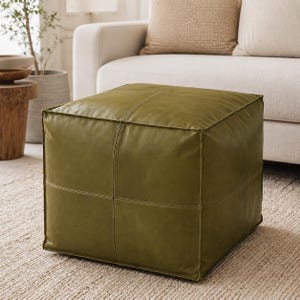 Op de afbeelding: Een vierkante olijfgroene lederen poef met zichtbare stiknaden die elke zijde in vier panelen verdelen. De poef heeft een gladde, matte afwerking en een gestructureerde, hoekige vorm, rustend op een lichtgekleurd geweven tapijt.