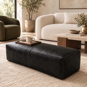 May include: A rectangular black leather ottoman featuring visible white stitching along the seams. A wooden tray sits on top of the ottoman, holding a small ceramic bowl and a light-colored ceramic mug. The ottoman has a smooth, matte finish.