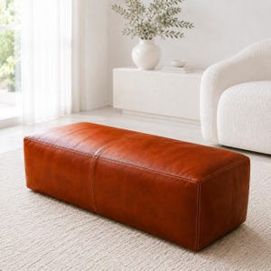 Puede incluir: Un otomano rectangular tapizado en cuero liso de color coñac. La pieza presenta una silueta limpia y cuadrada con costuras visibles que recorren el centro y los bordes, proporcionando una apariencia estructurada y uniforme.