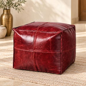 May include: A square, deep red leather pouf ottoman featuring a smooth, slightly distressed texture with visible stitched seams forming a grid pattern across its top and sides. The piece has a structured, boxy shape with defined edges and corners.