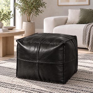 May include: A square, black leather ottoman featuring a structured, cube-like shape with visible stitched seams forming a grid pattern across its top and sides. The surface has a smooth, slightly reflective texture with subtle natural grain details.