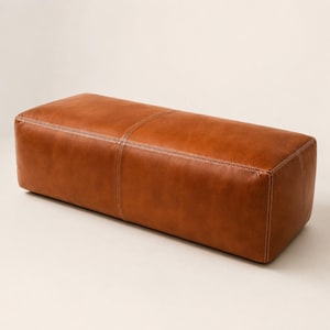 May include: A rectangular ottoman upholstered in smooth, cognac-colored leather. The piece features a central vertical seam and horizontal stitching along the top edges, showcasing a clean, boxy silhouette with visible light-colored thread detailing.