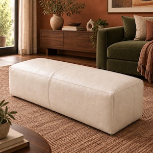May include: A rectangular, off-white leather ottoman with a smooth, matte finish and a central vertical seam. The piece features a low-profile, boxy silhouette with clean, straight edges and visible stitching along the top and side panels.