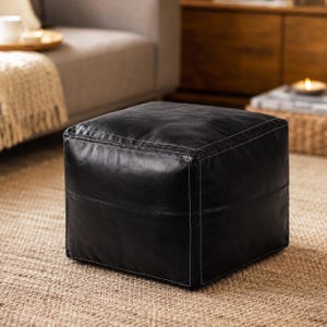 Pode incluir: Um pufe quadrado de couro preto com costuras brancas visíveis ao longo das bordas e junções. A superfície do pufe tem um acabamento liso e levemente texturizado com um brilho fosco, formando uma estrutura cúbica sólida.