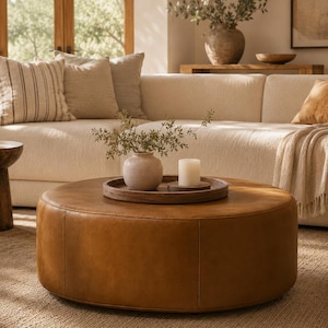 Puede incluir: Un otomano redondo de cuero color canela sirve como mesa de centro, cubierto con una bandeja circular de madera que sostiene un pequeño jarrón de cerámica clara con ramas verdes y una vela de pilar blanca sobre una base de madera, creando un punto focal.