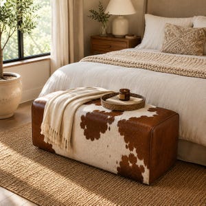 May include: A rectangular ottoman upholstered in brown and white cowhide featuring a smooth, low-profile design. A cream-colored fringed throw blanket is draped over one end, and a round woven tray holds a small candle and a book on top.