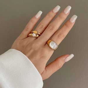 May include: A hand displaying three gold rings. One ring features a pearl, and another has a white, square-shaped stone. The rings are set against a neutral backdrop, with a white sleeve visible.